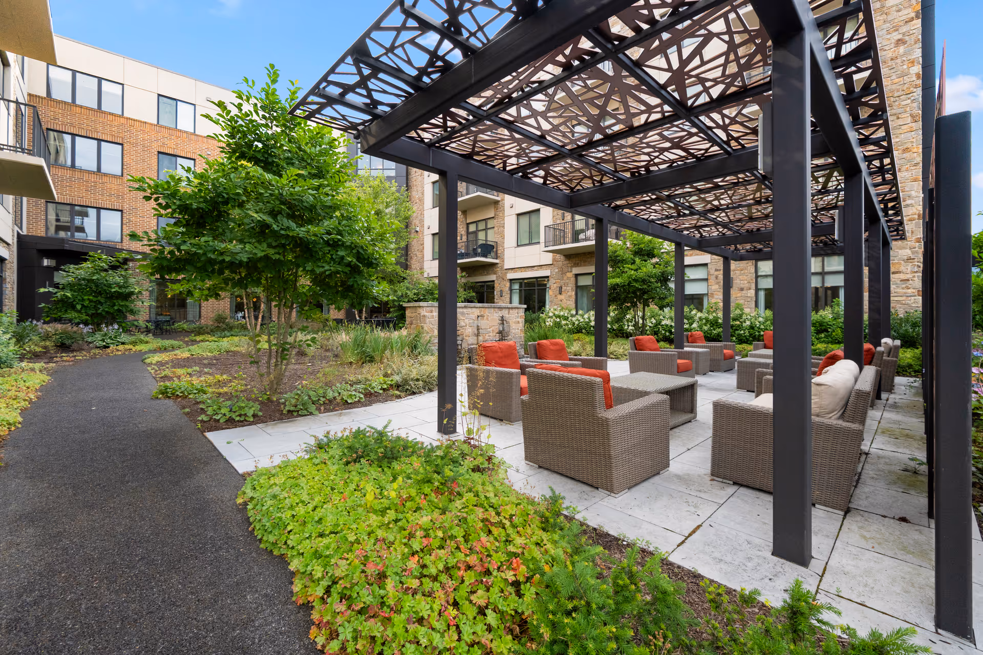 Outdoor seating area at Eagleview Landing with wicker chairs featuring red and beige cushions arranged under a decorative metal pergola. The area is surrounded by greenery, including bushes and trees, with a paved walkway and multi-story building in the background.