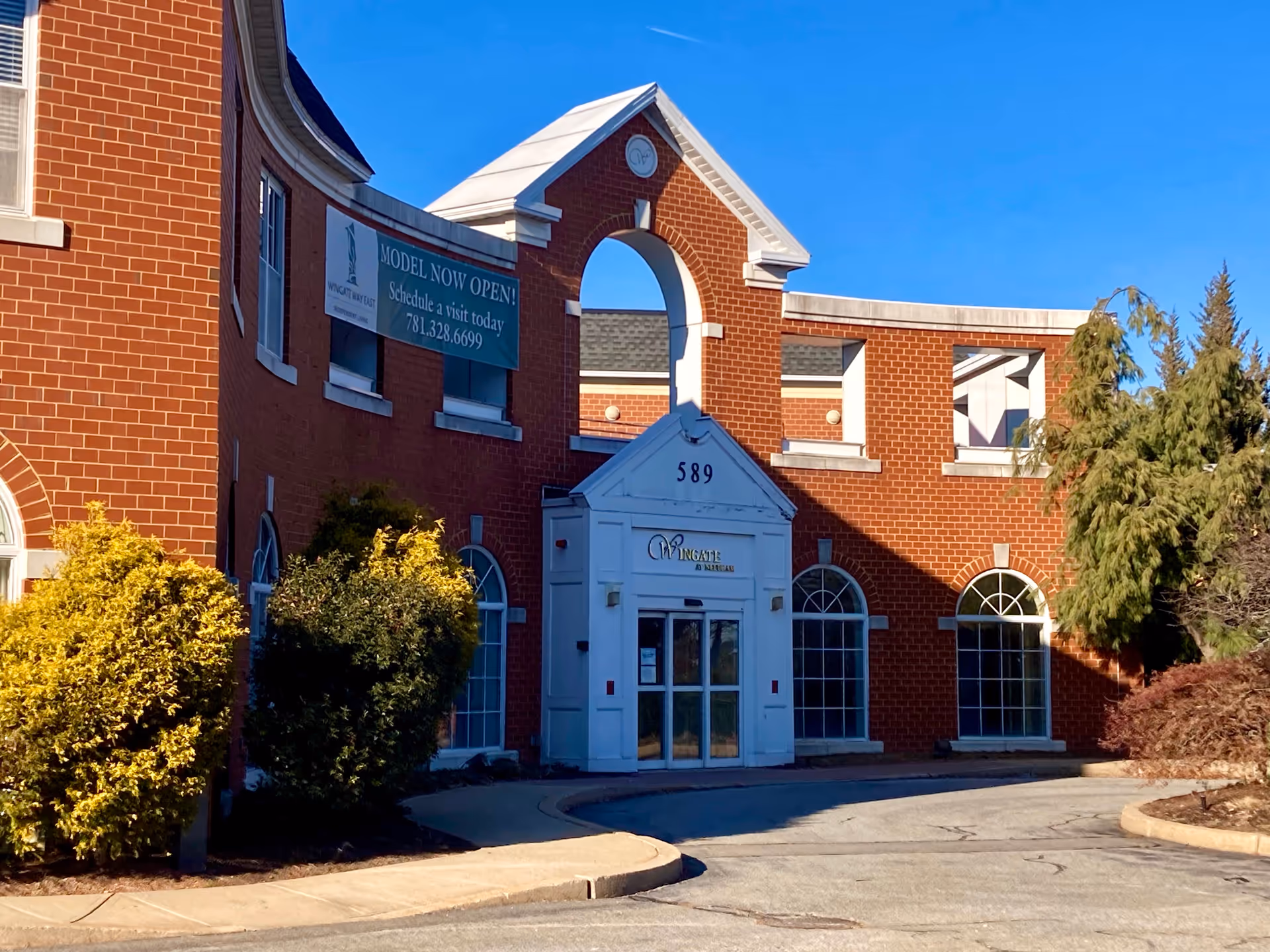 Front exterior view of a red brick building with white trim and arched windows, featuring an entrance with the sign 'Wingate At Needham' and the number 589 above the door. There is a green banner on the building that reads 'MODEL NOW OPEN! Schedule a visit today 781.328.6699'. Shrubs and trees surround the entrance under a clear blue sky.