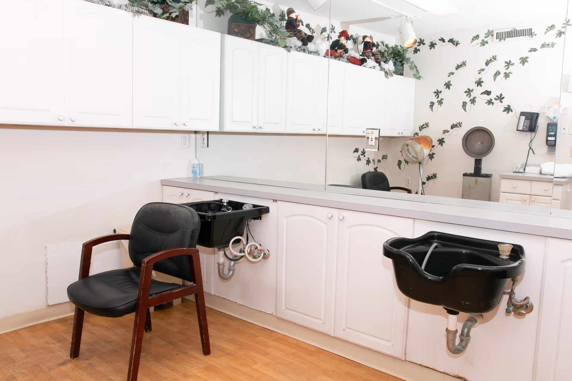 Interior of a hair salon area in a healthcare facility with two black salon sinks, a black salon chair with wooden armrests, white cabinets above and below a long countertop, a large mirror on the wall, and decorative greenery and figurines on top of the cabinets. The wall has a leafy vine pattern, and there is a vintage hair dryer in the corner.