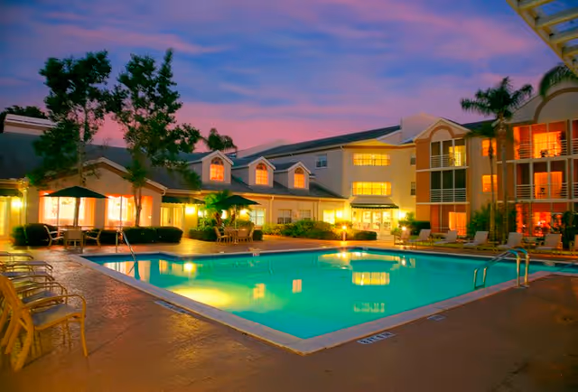 Outdoor swimming pool area at dusk with illuminated water, surrounded by chairs and tables with umbrellas. The pool is adjacent to a multi-story building with lit windows and palm trees around the area.