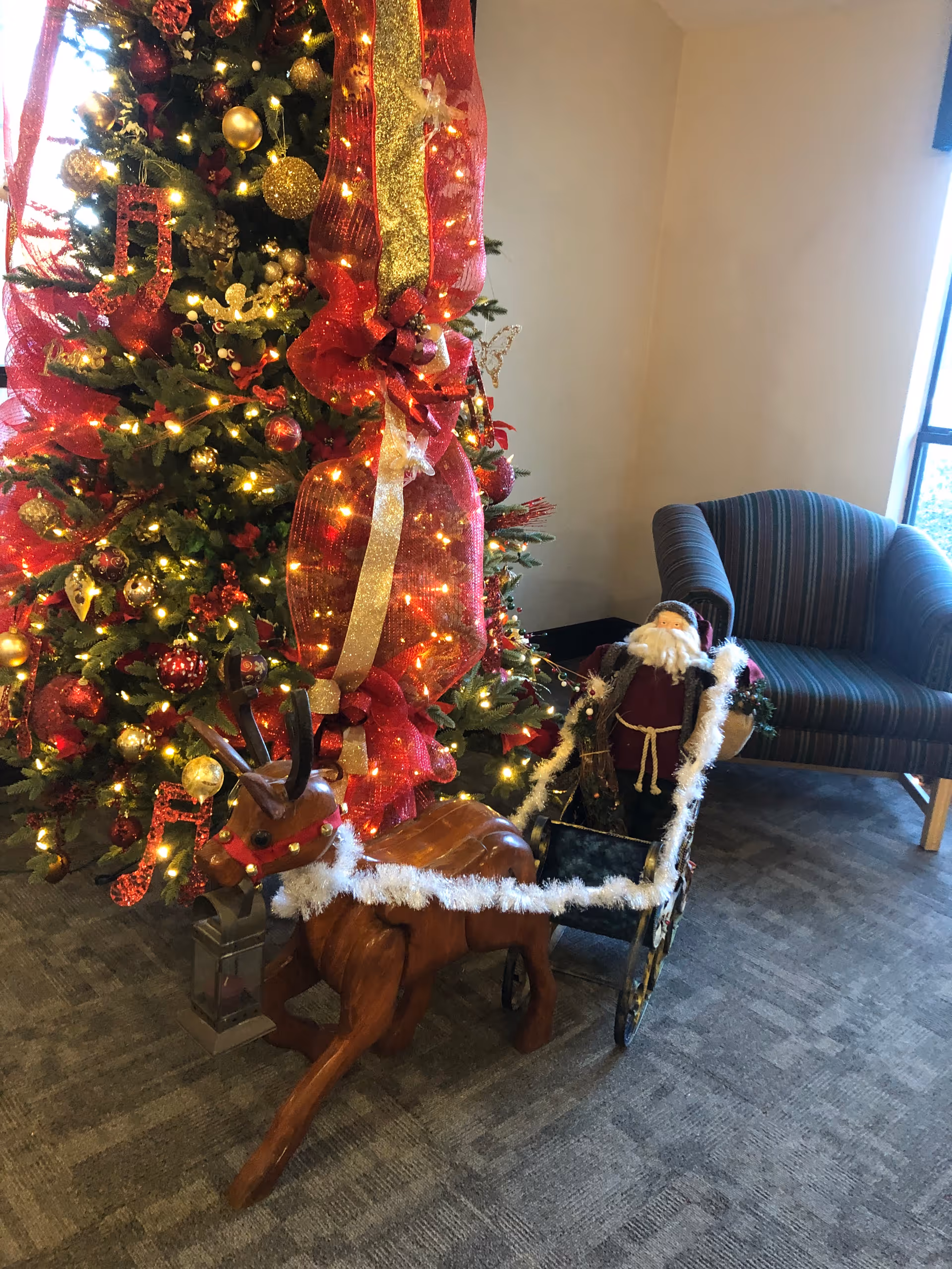 A decorated Christmas tree with red and gold ornaments and ribbons, next to a wooden reindeer pulling a small sleigh with a Santa Claus figure inside. A striped armchair is positioned near a window in the background.