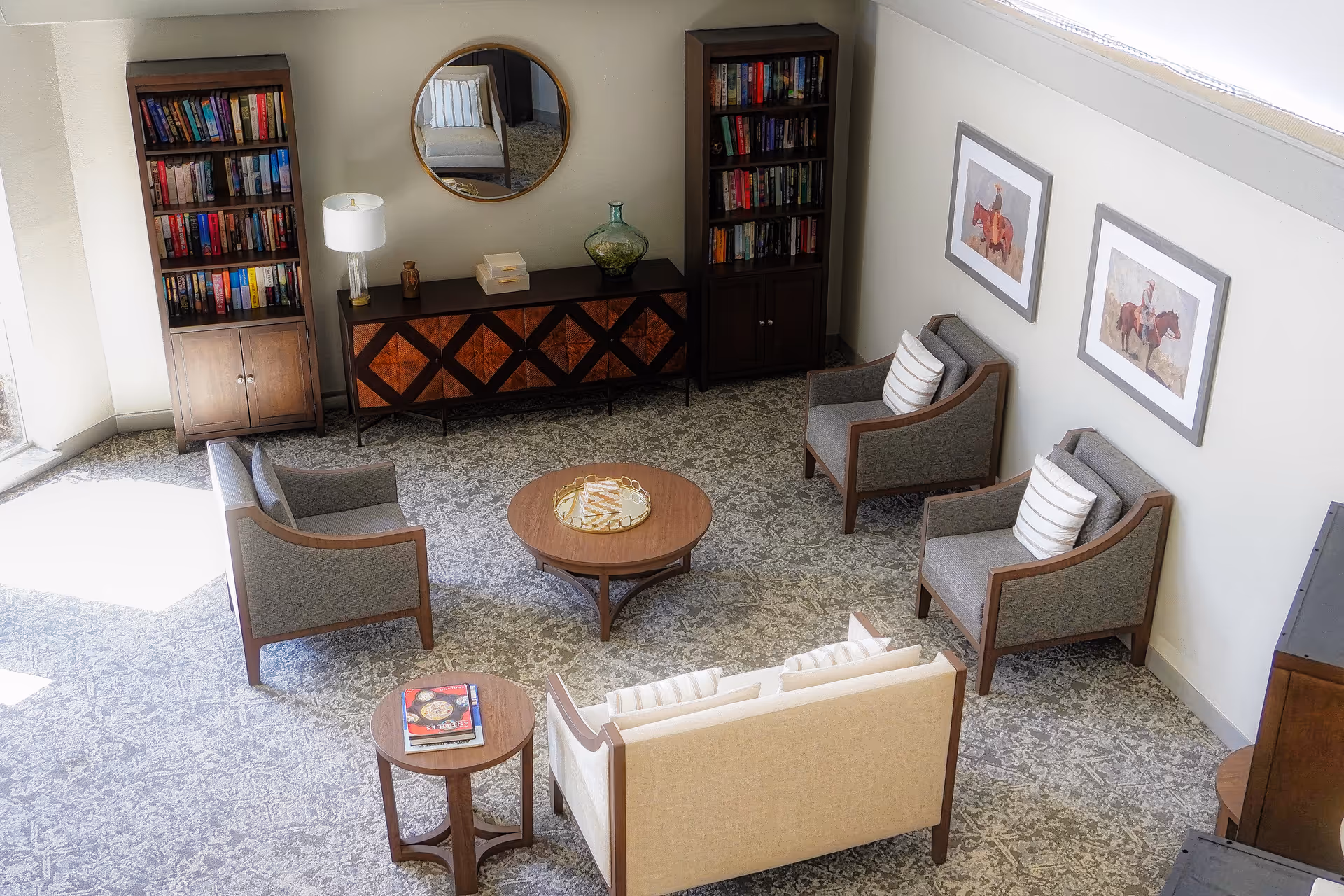 A cozy living room area with a beige sofa, three gray armchairs with striped cushions, a round wooden coffee table with a decorative tray, and a smaller round side table with books. Two tall wooden bookshelves filled with books flank a wooden cabinet with a decorative vase, lamp, and books on top. A round mirror hangs above the cabinet, and two framed pictures of people on horses are on the wall. The room has a patterned carpet and is well-lit by natural light from a large window.