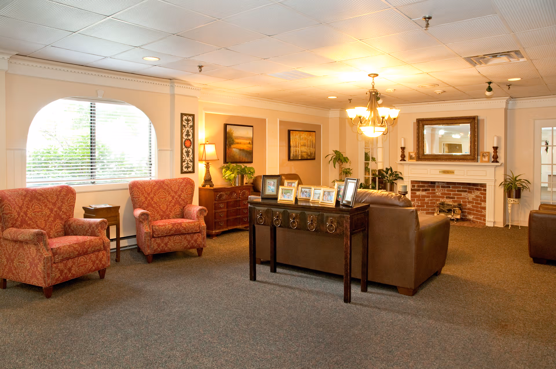 A cozy living room area with two red patterned armchairs near a window with blinds, a wooden side table with a lamp, a dark brown leather sofa, and a wooden console table displaying several framed photos. The room features a brick fireplace with a large mirror above it, plants, and warm lighting from a chandelier and lamps.