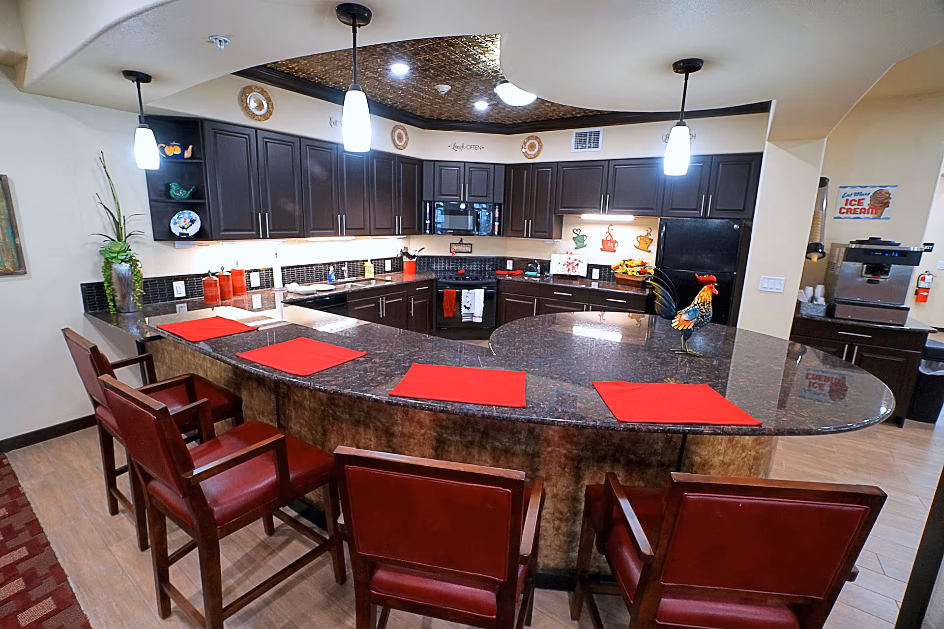 A modern kitchen area with dark wooden cabinets, a curved granite countertop with red placemats, and four red cushioned chairs. The kitchen features pendant lights hanging from the ceiling, a black refrigerator, a microwave, and various decorative items including a rooster figurine and wall art. There is also an ice cream sign and a coffee machine visible in the background.