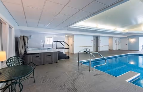 Indoor swimming pool area with a hot tub, metal handrails, and a small round table with two chairs. The space has a tiled floor, white walls, and a ceiling with recessed lighting.