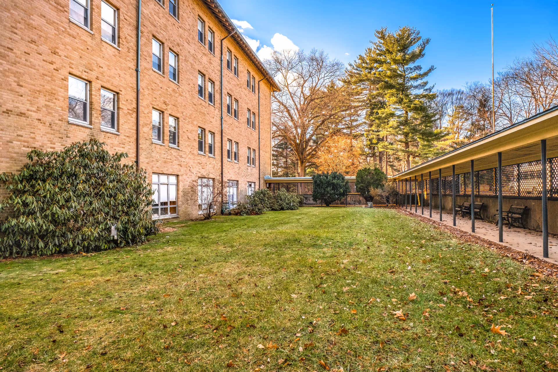 Outdoor grassy courtyard area at Monastery Heights Senior Living & Memory Care with a large brick building on the left and a covered walkway with benches on the right. Trees and shrubs surround the space under a blue sky with some clouds.