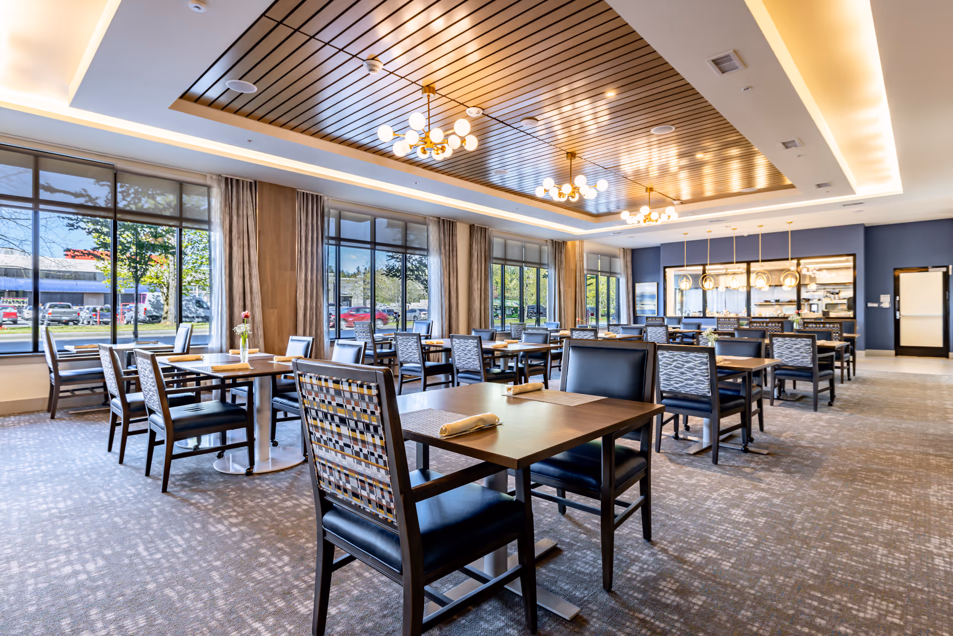 A spacious and well-lit dining room with multiple wooden tables and chairs arranged neatly. Large windows along one wall allow natural light to fill the room, showing a view of the outside greenery and parked cars. The ceiling features modern wooden paneling with elegant hanging light fixtures. The room has a carpeted floor and a serving area with pendant lights in the background.