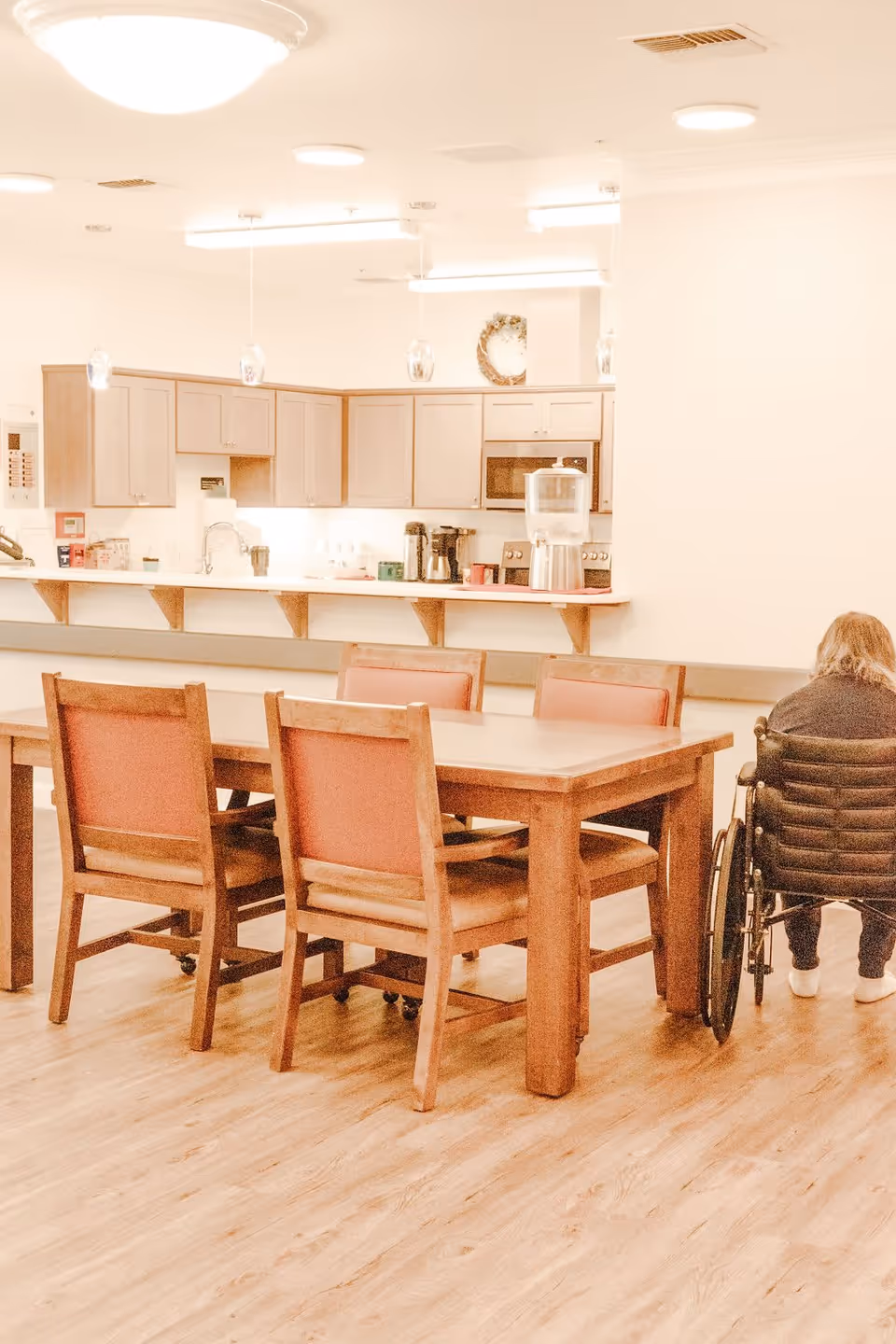 Dining area with a wooden table and chairs in front of a kitchenette, and a person in a wheelchair seated on the right.