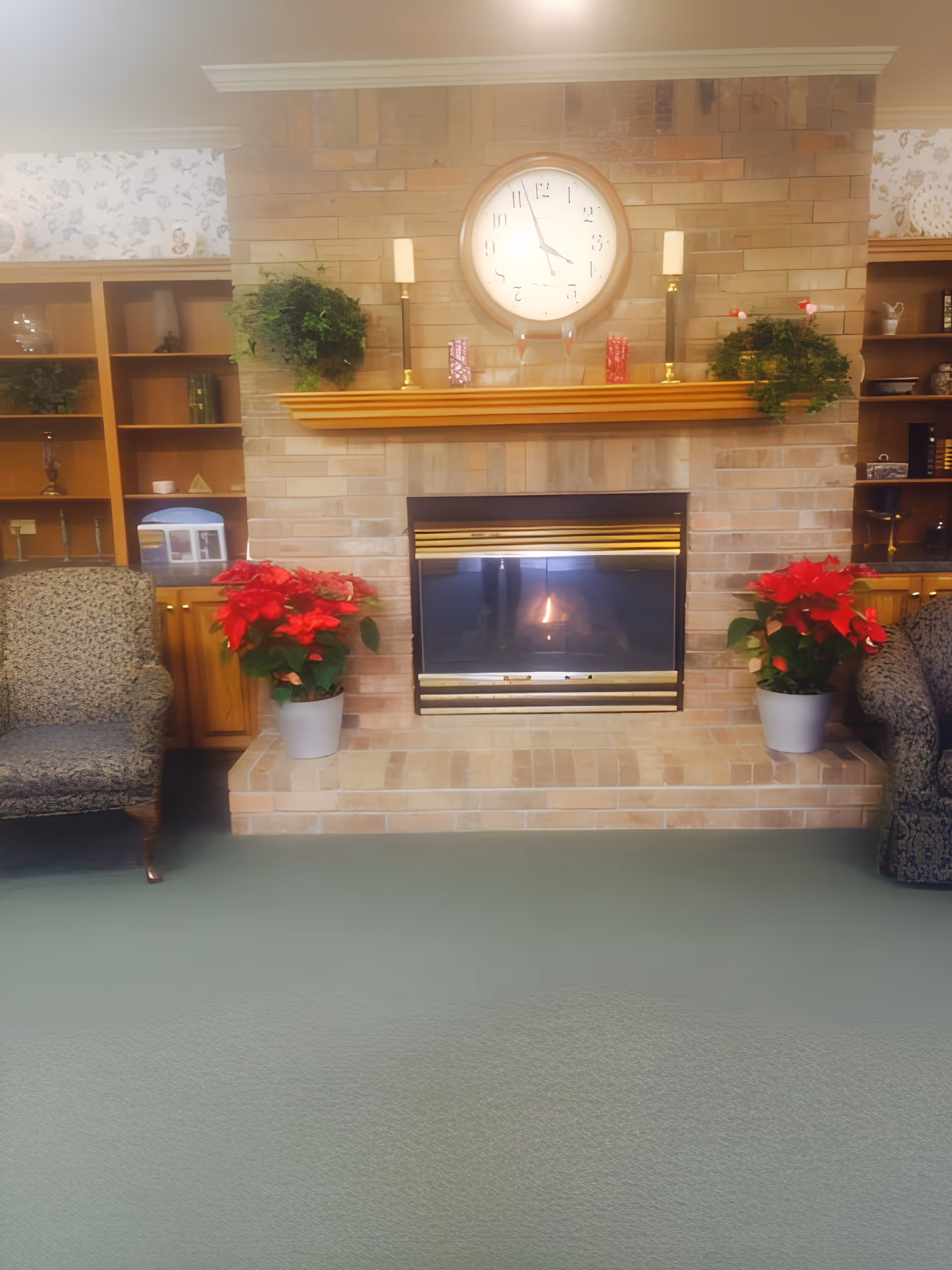 A cozy living room area featuring a brick fireplace with a fire burning inside. Above the fireplace is a large round clock and two tall candle holders on a wooden mantel decorated with green plants and red decorative items. On either side of the fireplace are built-in wooden shelves with various decorative objects. Two patterned armchairs flank the fireplace, and two potted poinsettia plants are placed on the hearth.