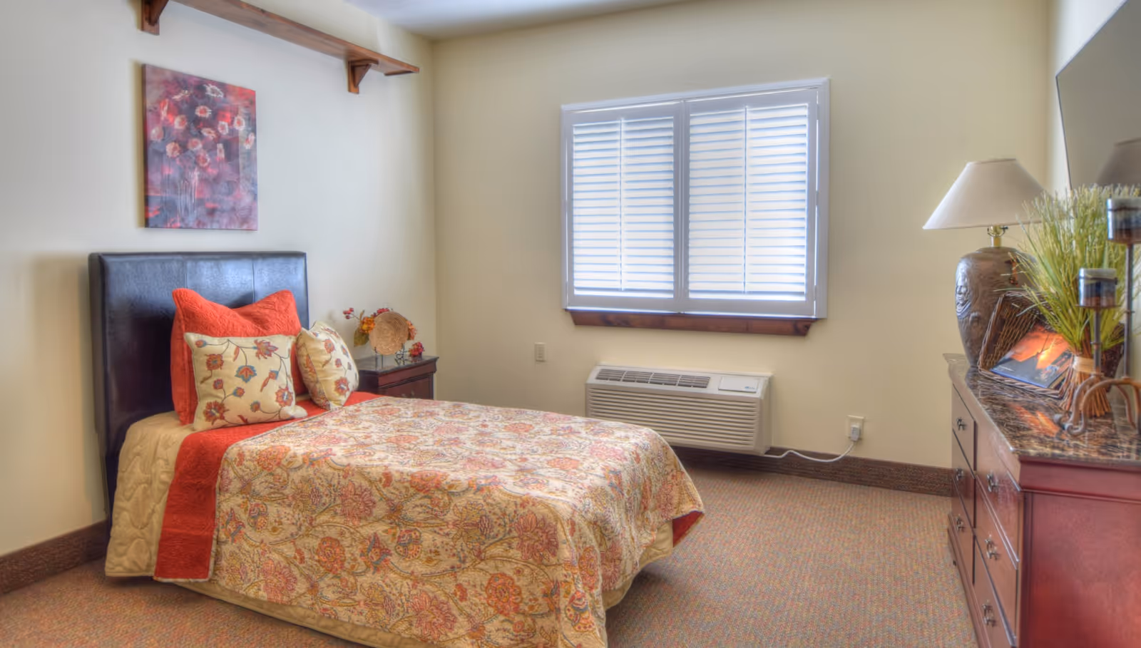 A cozy single bedroom with a patterned bed, decorative pillows, nightstand, window with blinds, and a dresser with a lamp and decor.