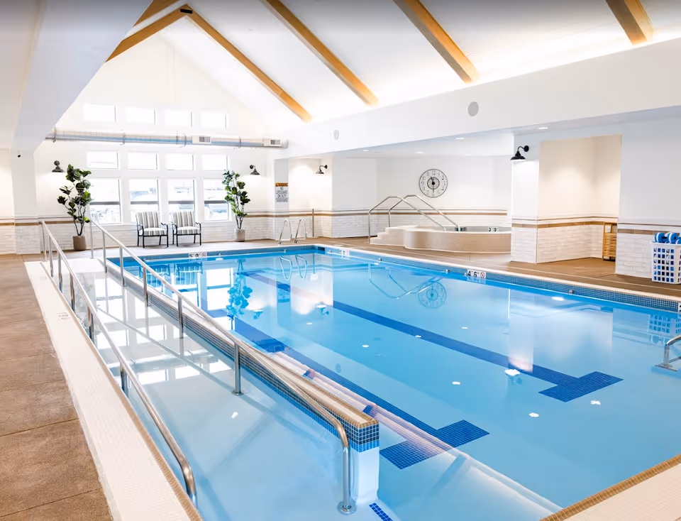 Indoor swimming pool area with a ramp for accessibility, two chairs near large windows, potted plants, a hot tub in the corner, and a clock on the wall. The ceiling has wooden beams and bright lighting.