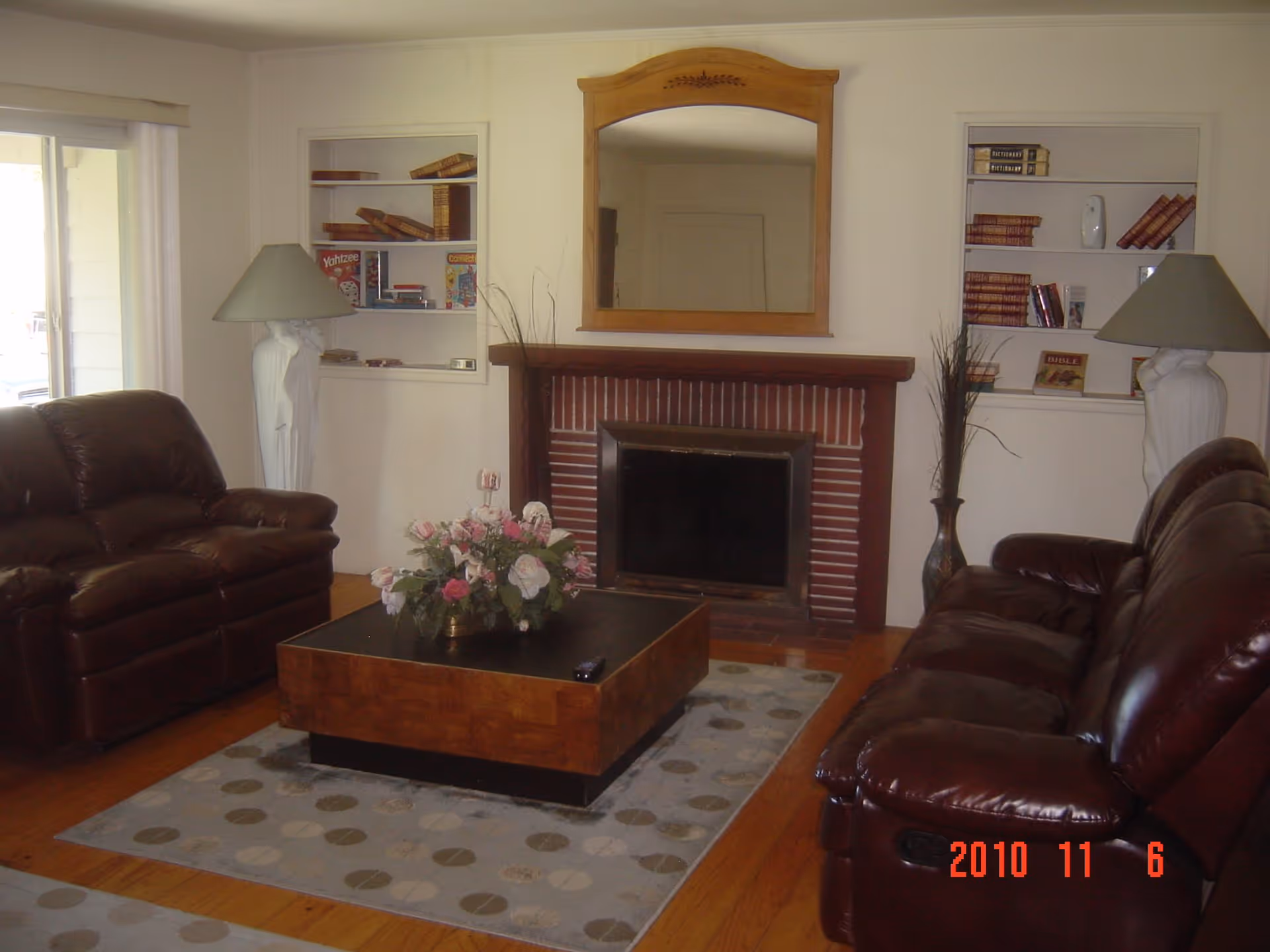 A cozy living room with two dark brown leather sofas facing a brick fireplace with a wooden mantel and a large mirror above it. There is a square wooden coffee table with a floral arrangement in the center on a patterned rug. Built-in shelves on either side of the fireplace hold books, games, and decorative items. Two white lamps with beige shades stand on either side of the fireplace.