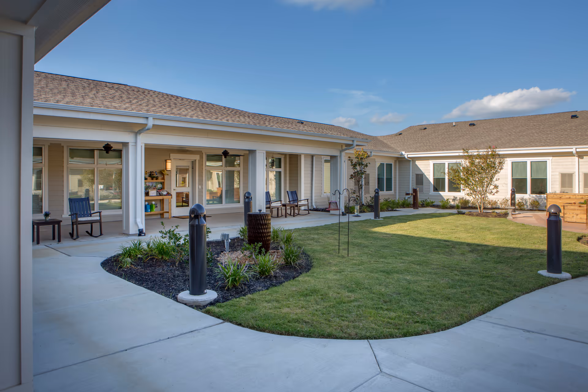 Outdoor courtyard area of a senior living facility with a well-maintained lawn, small garden beds, and a concrete walkway. The building surrounds the courtyard with large windows and a covered patio area furnished with rocking chairs and small tables. The sky is clear with a few clouds.