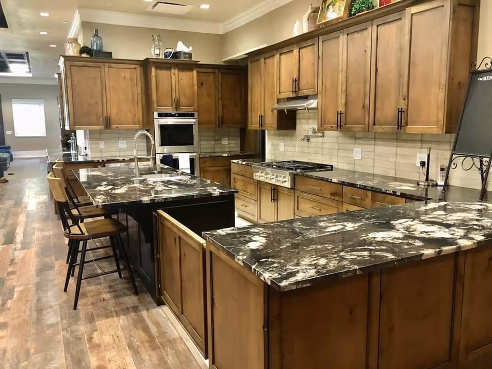A spacious kitchen with wooden cabinets and dark marble countertops. The kitchen features a large island with a sink and three wooden bar stools. There is a built-in oven, a stovetop with a range hood, and a tiled backsplash. The floor is wooden, and the kitchen opens into a hallway with natural light coming from a window in the background.