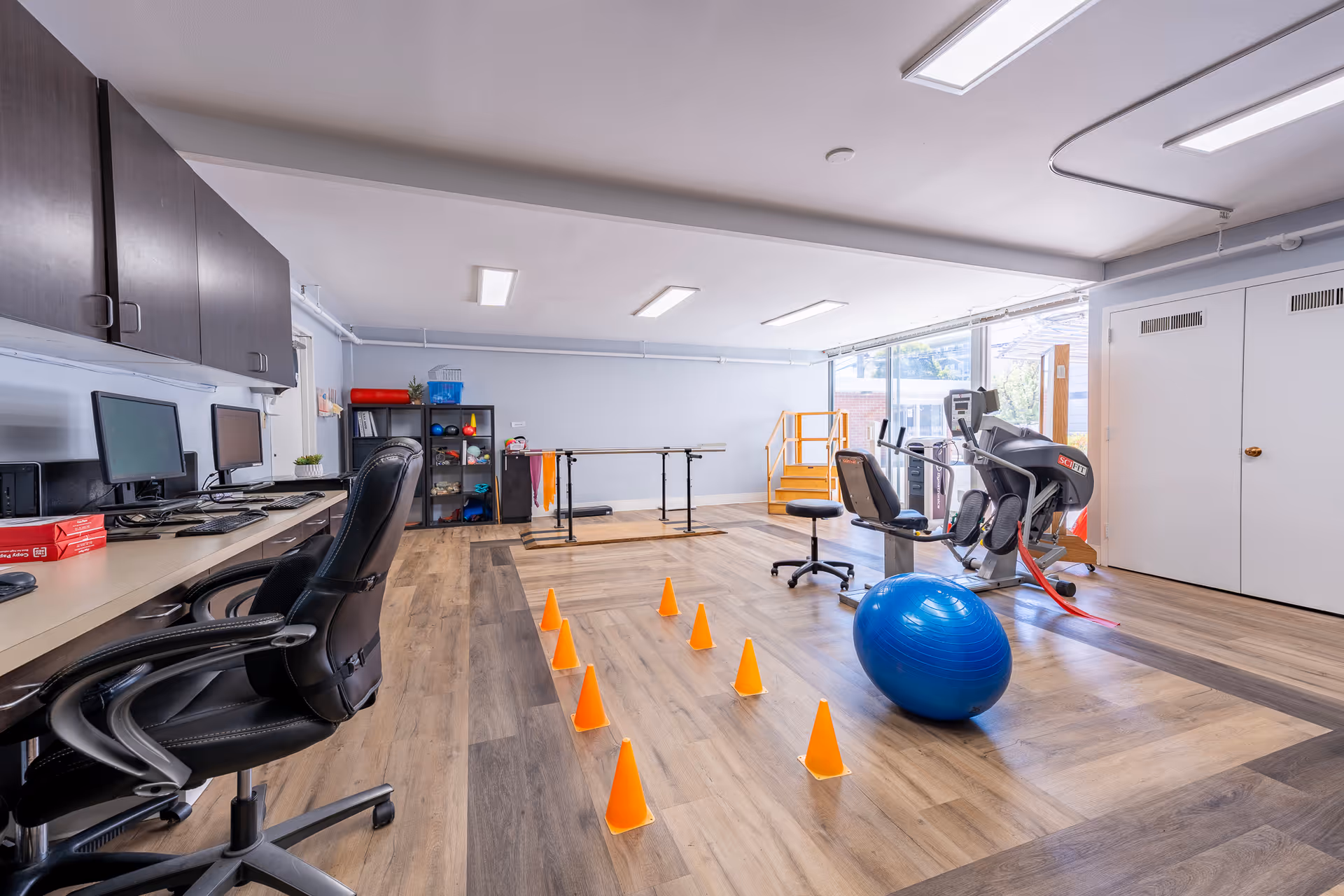 Bright interior therapy and fitness room with desks and computers, orange training cones and a blue stability ball, and cardio equipment by a window.