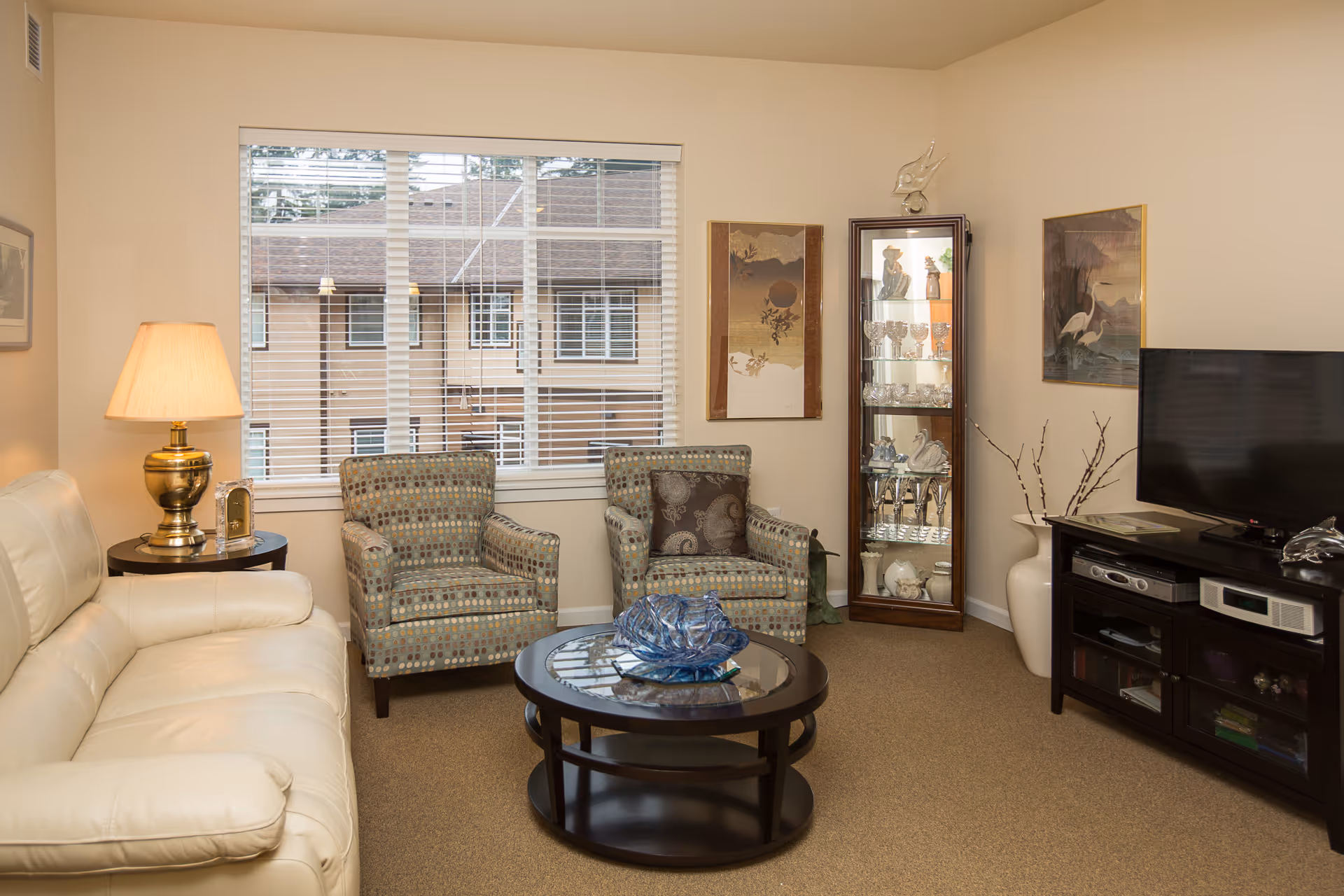 A cozy living room with a cream leather sofa, two patterned armchairs, a round glass coffee table with a decorative blue glass piece, a wooden TV stand with a flat-screen TV, and a tall glass display cabinet filled with decorative items. The room has beige walls, a large window with white blinds, and framed artwork on the walls.