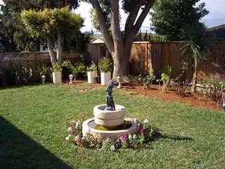 Outdoor backyard with a statue fountain, circular planter, flowers, and green lawn.