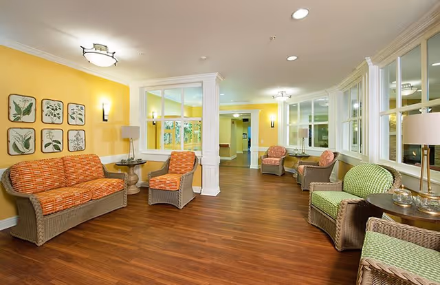 A bright and spacious common area with yellow walls and wood flooring, furnished with wicker chairs and sofas featuring orange and green cushions. The room has large windows, decorative wall art with botanical prints, and several table lamps providing warm lighting.