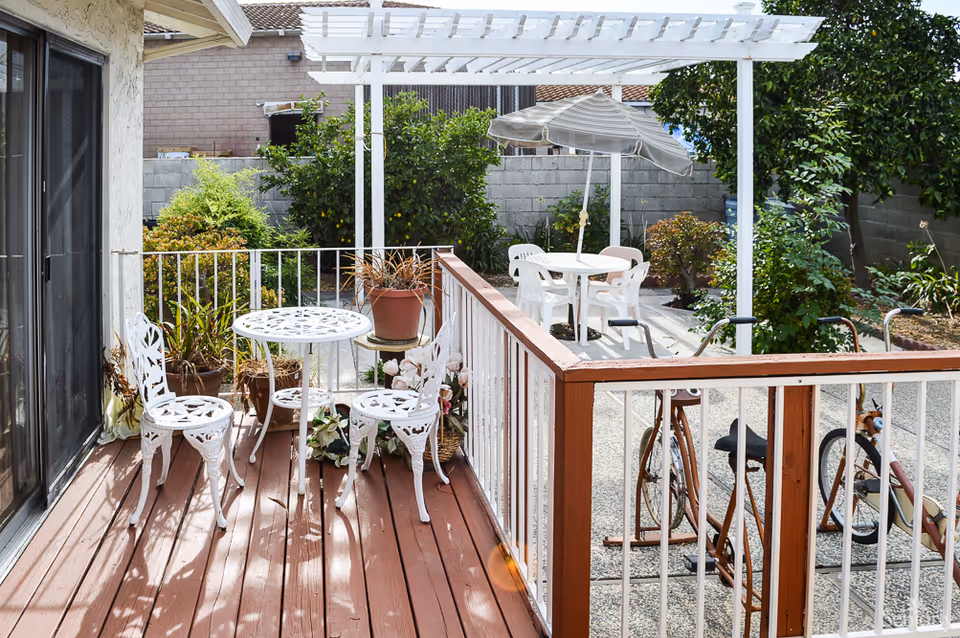 Outdoor patio area with a small white metal table and three matching chairs on a wooden deck. There are potted plants around the seating area. Beyond the deck, there is a white pergola covering a concrete patio with a round table, four white plastic chairs, and a large striped umbrella. The area is surrounded by greenery and a gray concrete block wall.