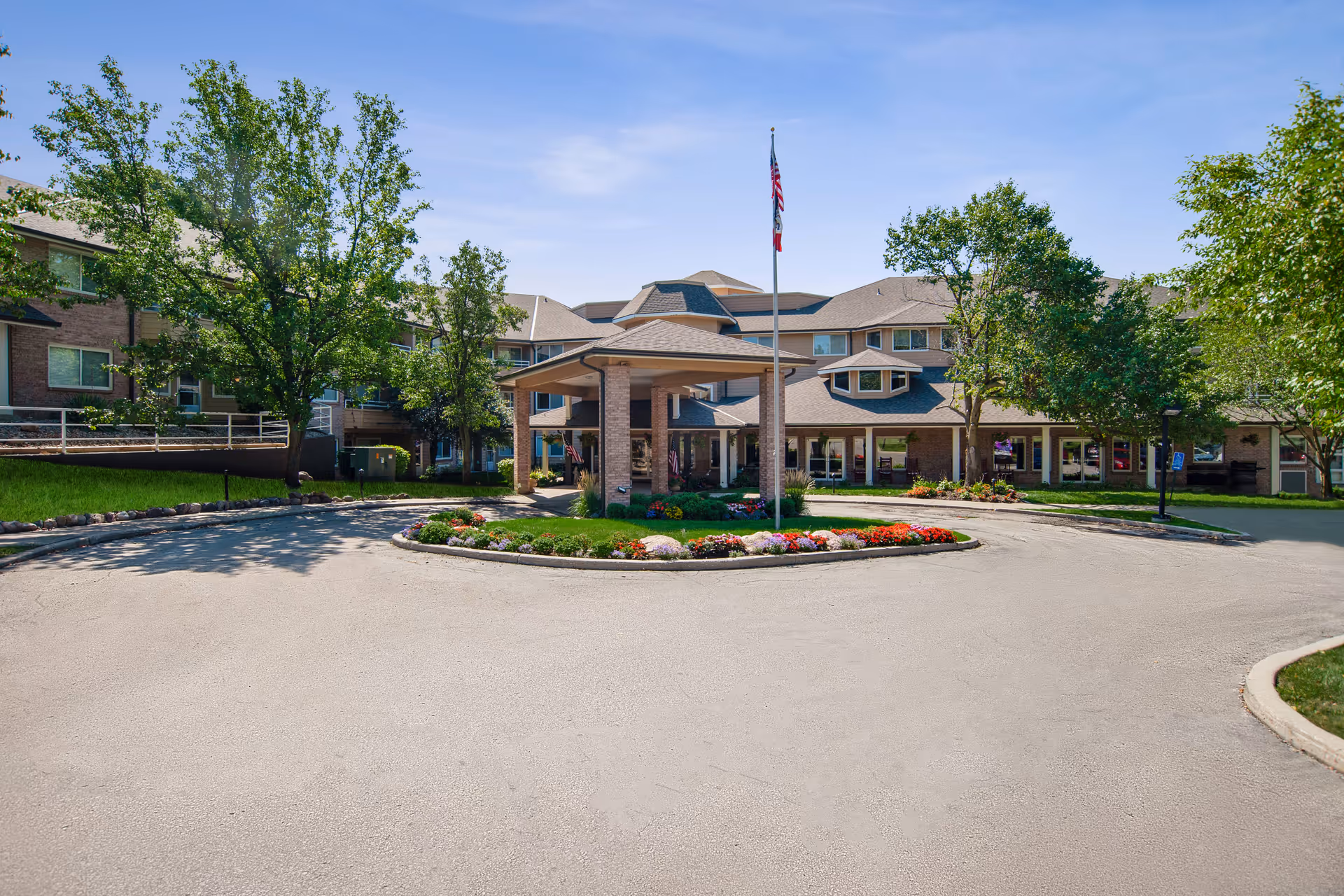 Front exterior view of Independence Village of Des Moines, showing a circular driveway with a landscaped island in the center featuring colorful flowers and a flagpole with an American flag. The building is a multi-story structure with large windows and surrounded by trees under a clear blue sky.