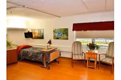 A cozy bedroom in a senior living facility with a single bed covered in a colorful quilt, a small wall-mounted TV, two chairs with a small table between them, and two framed pictures on the wall. The room has wooden flooring and a window with a red valance.