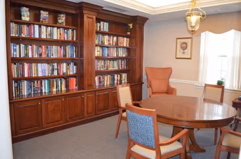 A cozy room with a large wooden bookshelf filled with books along one wall, a round wooden table surrounded by four chairs, and an upholstered armchair near a window with curtains. A framed picture and a hanging light fixture are also visible.
