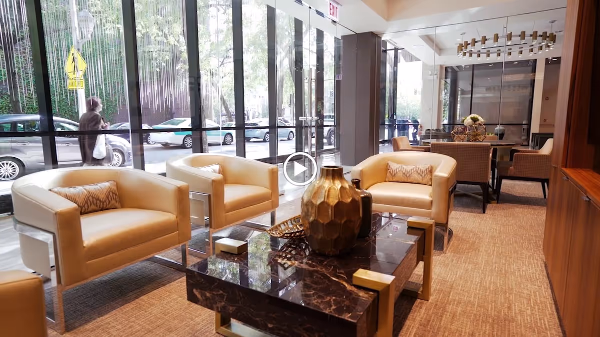 A modern and elegant lounge area with cream-colored armchairs arranged around a dark marble coffee table decorated with gold vases. Large floor-to-ceiling windows provide a view of parked cars and trees outside. The space features neutral tones, carpeted flooring, and a chandelier hanging from the ceiling.