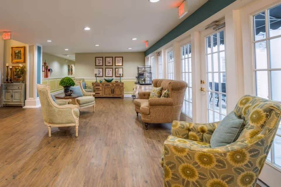 A bright and spacious lounge area in a senior living facility with wooden flooring, multiple upholstered armchairs with cushions, large windows and glass doors letting in natural light, and decorative furniture including a sideboard with framed artwork on the wall.