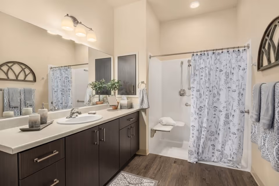 A modern bathroom with a large mirror above a dark wood vanity with a white countertop and sink. There are three light fixtures above the mirror. To the right is a walk-in shower with a white floral shower curtain, a fold-down seat, and grab bars. The bathroom has light-colored walls, wood-look flooring, and neatly folded towels on a rack.