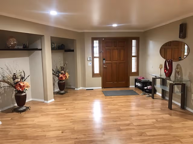 An entryway with a wooden front door flanked by two narrow windows. The floor is light wood laminate. To the left are two alcoves with decorative vases containing artificial flowers. To the right is a glass console table with decorative items and a round mirror above it. A small shoe rack with shoes is near the door.