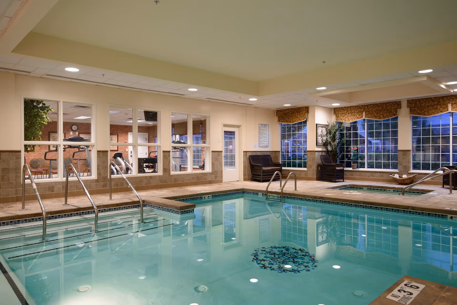 Indoor swimming pool area with clear water, tiled edges, and metal handrails. Adjacent to the pool is a hot tub with similar features. Large windows with patterned valances allow natural light to enter, and there are two wicker chairs and a potted plant near the windows. Through interior windows, a fitness room with exercise equipment and chairs is visible.