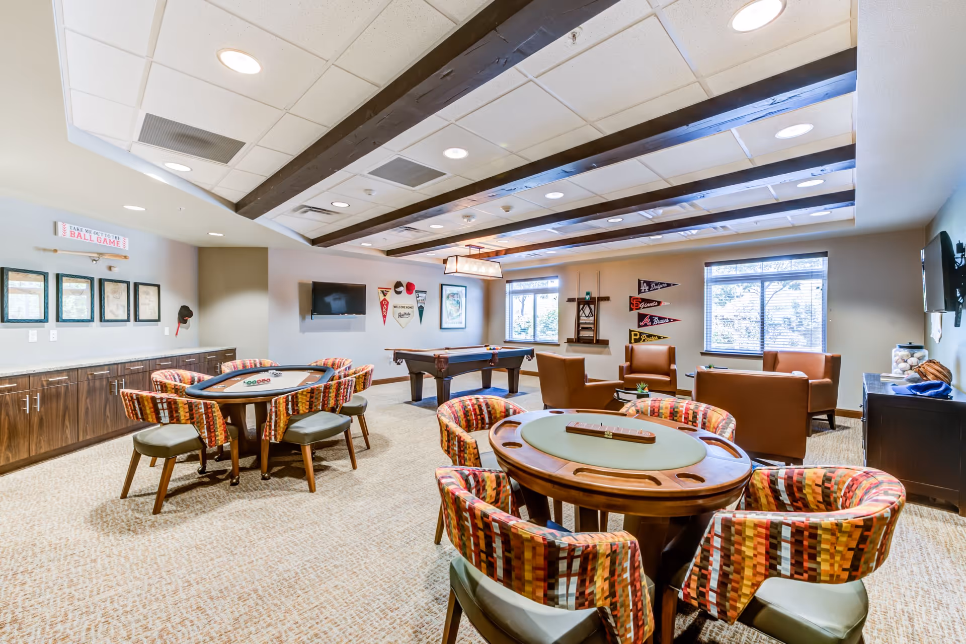 A bright and spacious game room with patterned chairs around two card tables, a pool table in the background, and a seating area with brown leather chairs and a sofa near two windows. The walls are decorated with sports pennants and framed pictures, and the ceiling features wooden beams with recessed lighting.