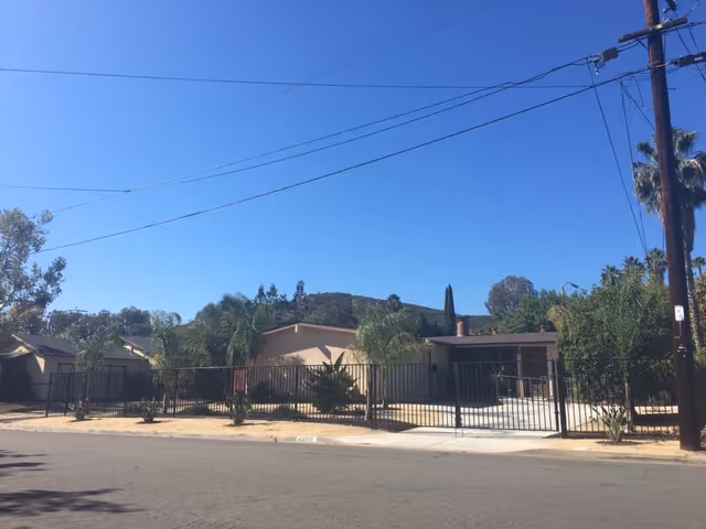 Exterior view of a single-story residential building with a fenced front yard, palm trees, and a clear blue sky. The building is situated on a paved street with utility poles and wires visible.
