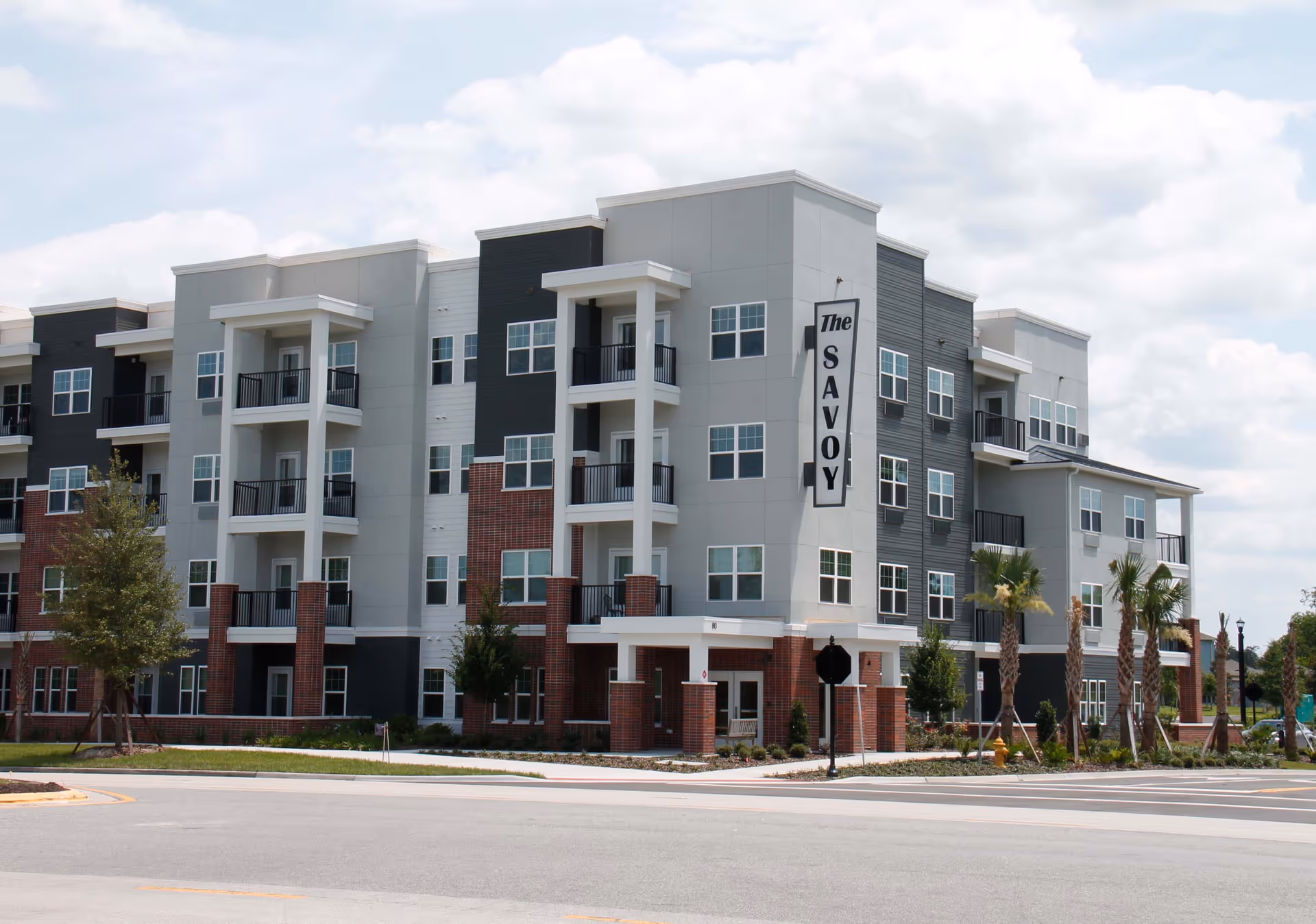 Exterior view of a modern multi-story residential building named The Savoy, featuring balconies, large windows, and a mix of gray, white, black, and brick facade under a partly cloudy sky.