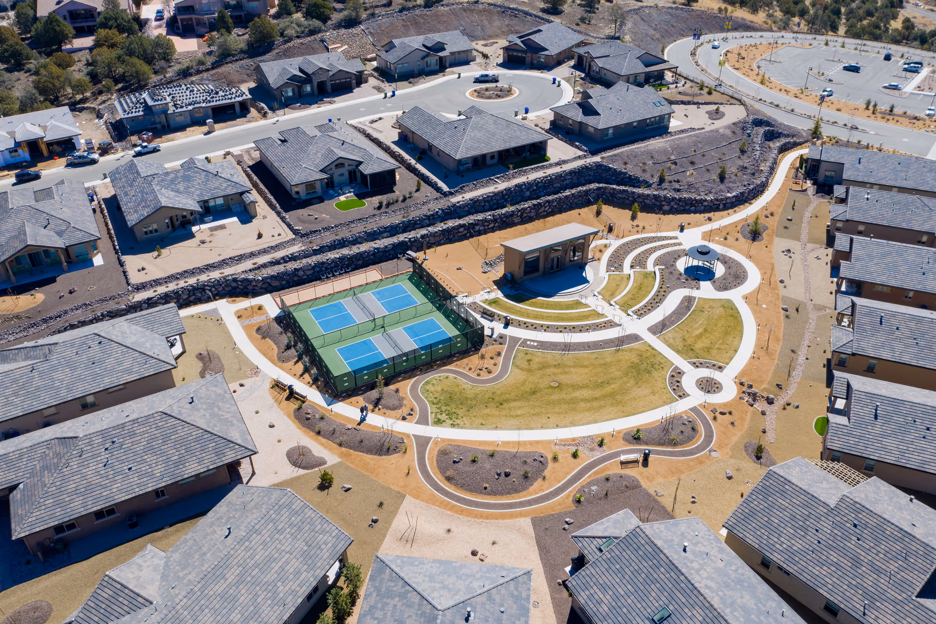Aerial view of a senior living community featuring multiple single-story residential buildings arranged around a central outdoor recreational area with tennis courts, walking paths, a small amphitheater, and landscaped green spaces.