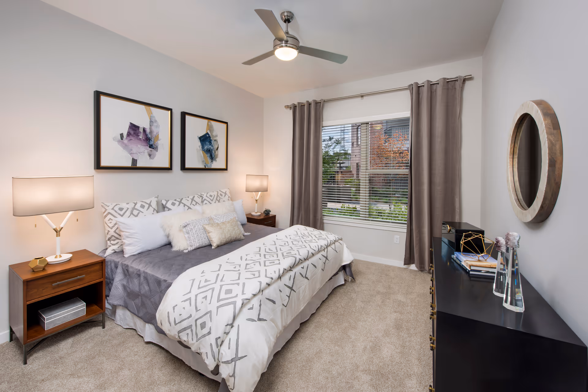 A neatly arranged bedroom with a bed featuring patterned bedding and multiple pillows. There are two framed abstract paintings above the bed. On either side of the bed are wooden nightstands with modern lamps. A window with brown curtains and blinds shows greenery outside. A black dresser with decorative items and a round wooden-framed mirror is on the right wall. The room has beige carpet and a ceiling fan with a light.