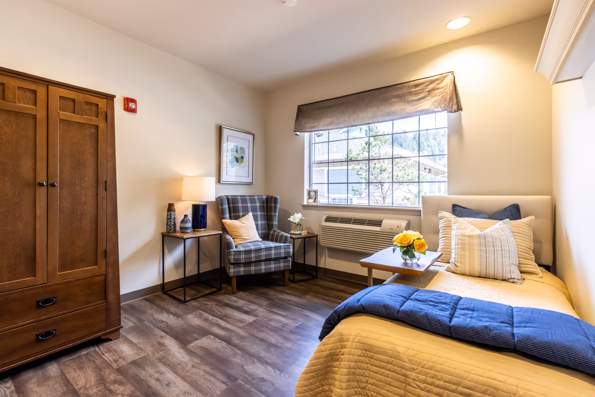 A cozy bedroom in a senior living facility with a wooden wardrobe, a plaid armchair with a pillow, two side tables with decorative items and a lamp, a large window with a view outside, and a bed with yellow bedding, multiple pillows, and a blue throw blanket. A small table with a vase of yellow flowers is placed on the bed.