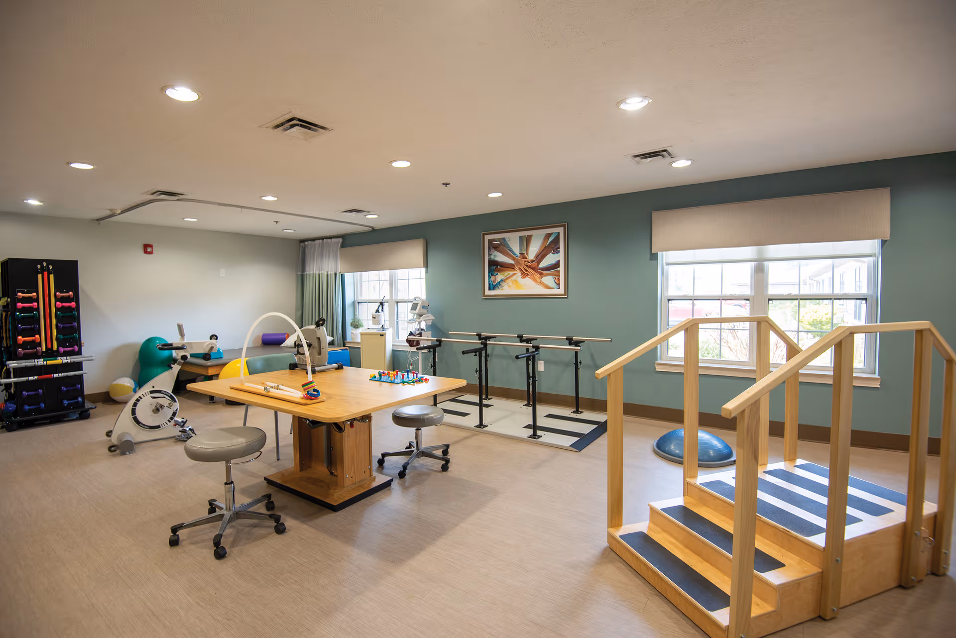 A bright rehabilitation therapy room with parallel bars, exercise stairs, a therapy table and assorted fitness equipment.