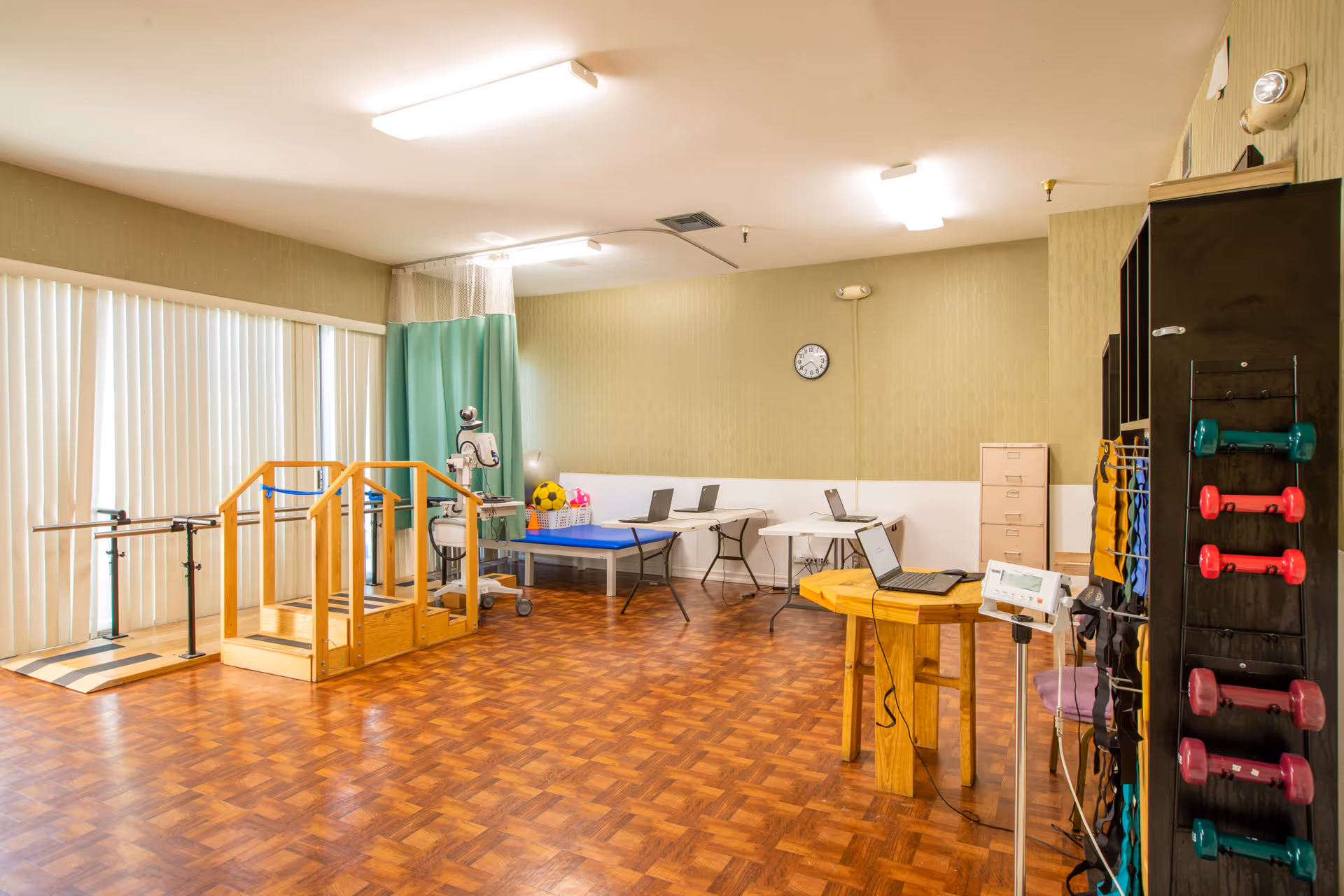 A spacious rehabilitation or physical therapy room with wooden flooring, parallel bars for walking exercises, a small set of wooden stairs, exercise equipment including colorful dumbbells on a rack, a treatment table, and several laptops on folding tables. The room has vertical blinds covering the windows and a clock on the wall.