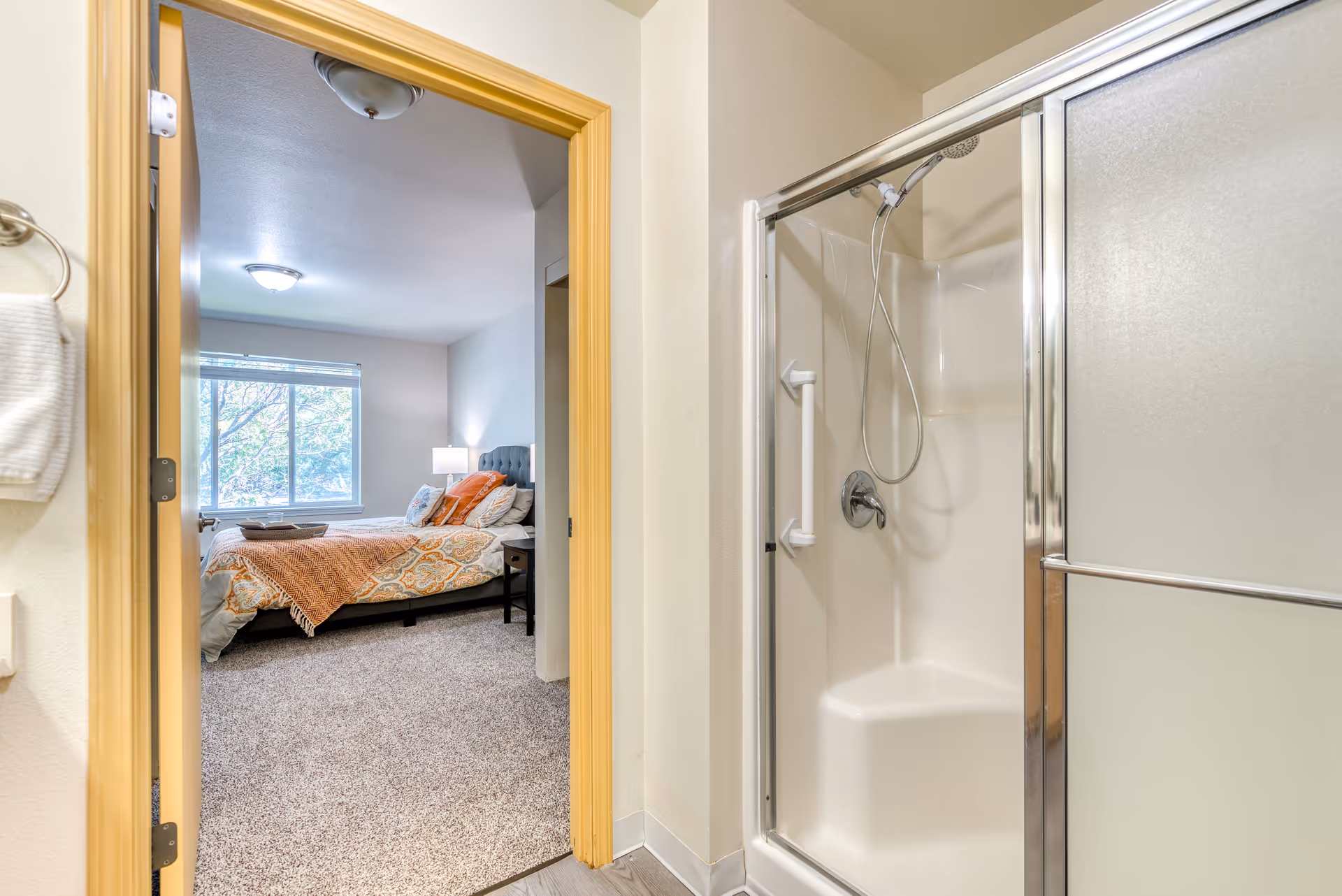 View from a bathroom showing a shower with a sliding glass door and a handheld showerhead. Through the open door, a bedroom is visible with a bed covered in patterned bedding and orange throw pillows, a nightstand with a lamp, and a large window letting in natural light.