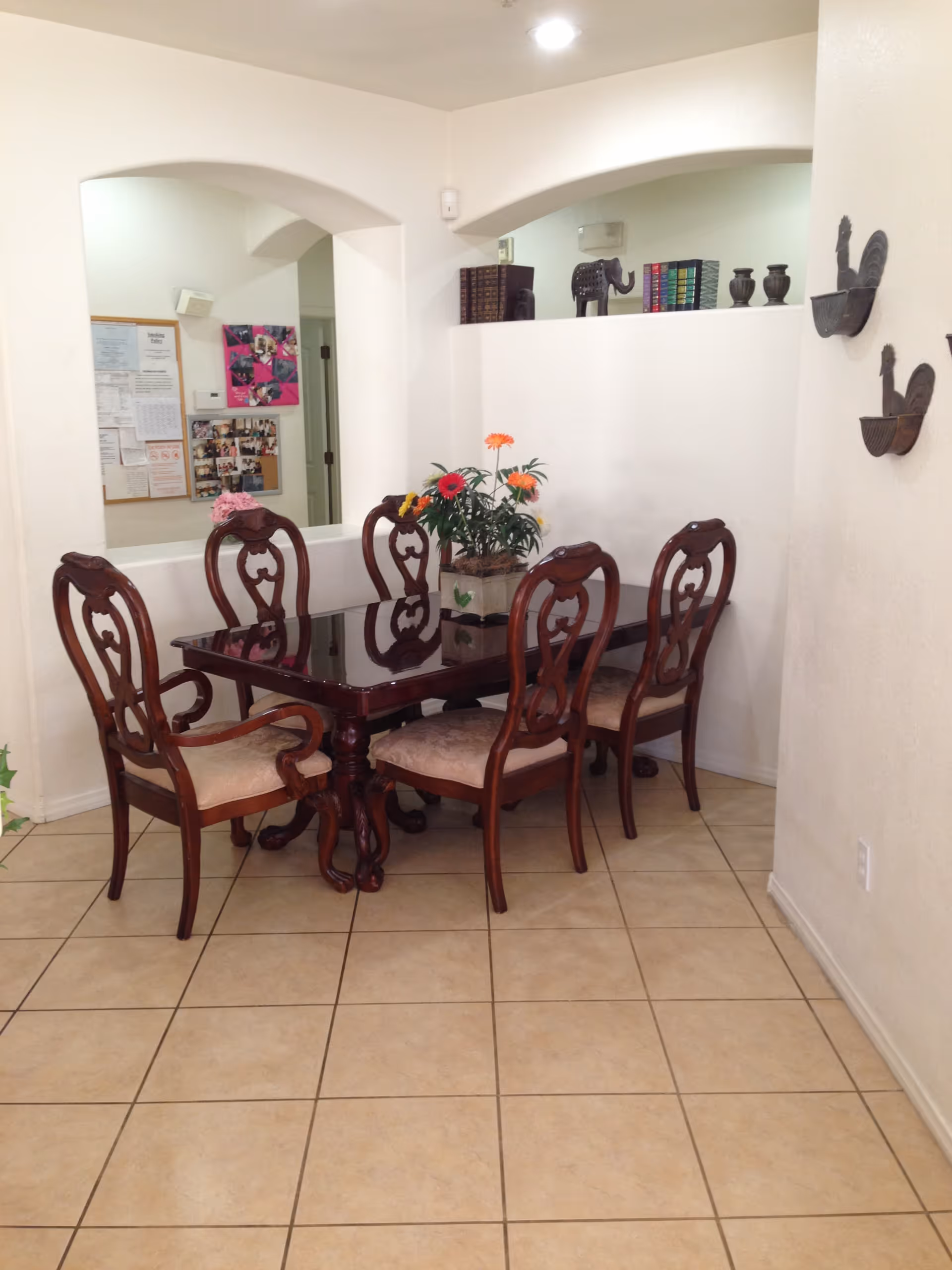 A dining area with a dark wooden table and six matching chairs with cushioned seats. A flower arrangement with colorful flowers is placed in the center of the table. The room has tiled flooring and white walls with decorative wall hangings shaped like roosters. There are two arched openings in the wall, one showing a bulletin board with papers and photos, and the other displaying decorative items including books and an elephant figurine.