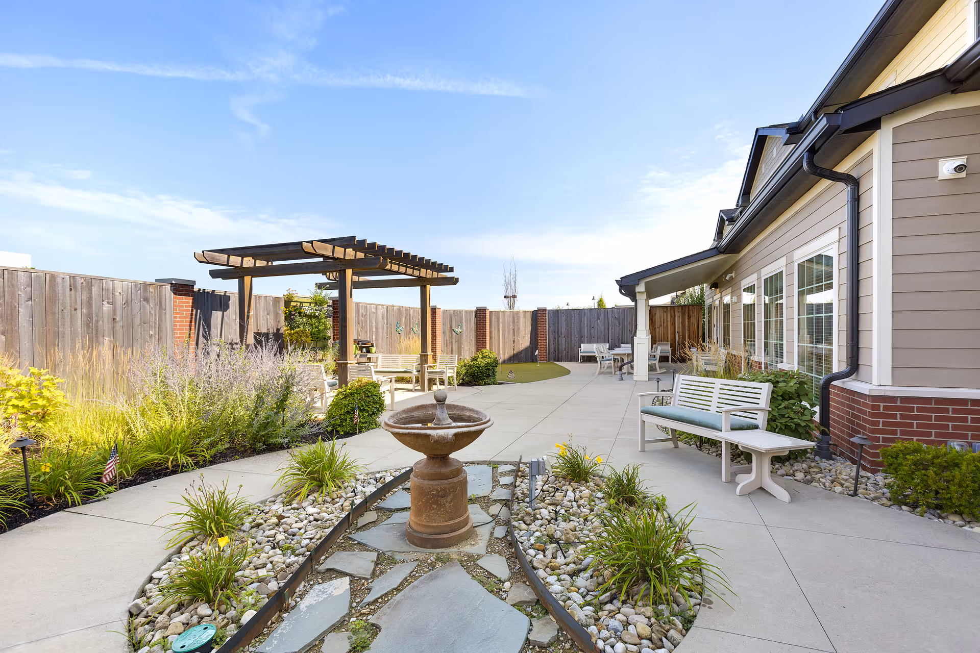 Outdoor patio area at StoryPoint Powell North featuring a paved walkway, a small water fountain surrounded by rocks and plants, benches with cushions, a wooden pergola with seating underneath, and a wooden privacy fence with brick pillars. The building exterior has beige siding with white trim and multiple windows.
