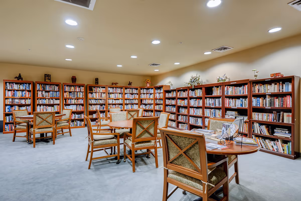 A spacious library room with multiple wooden bookshelves filled with books along the walls. Several round wooden tables with cushioned chairs are arranged in the center of the room. The room is well-lit with recessed ceiling lights and has a light-colored carpeted floor.