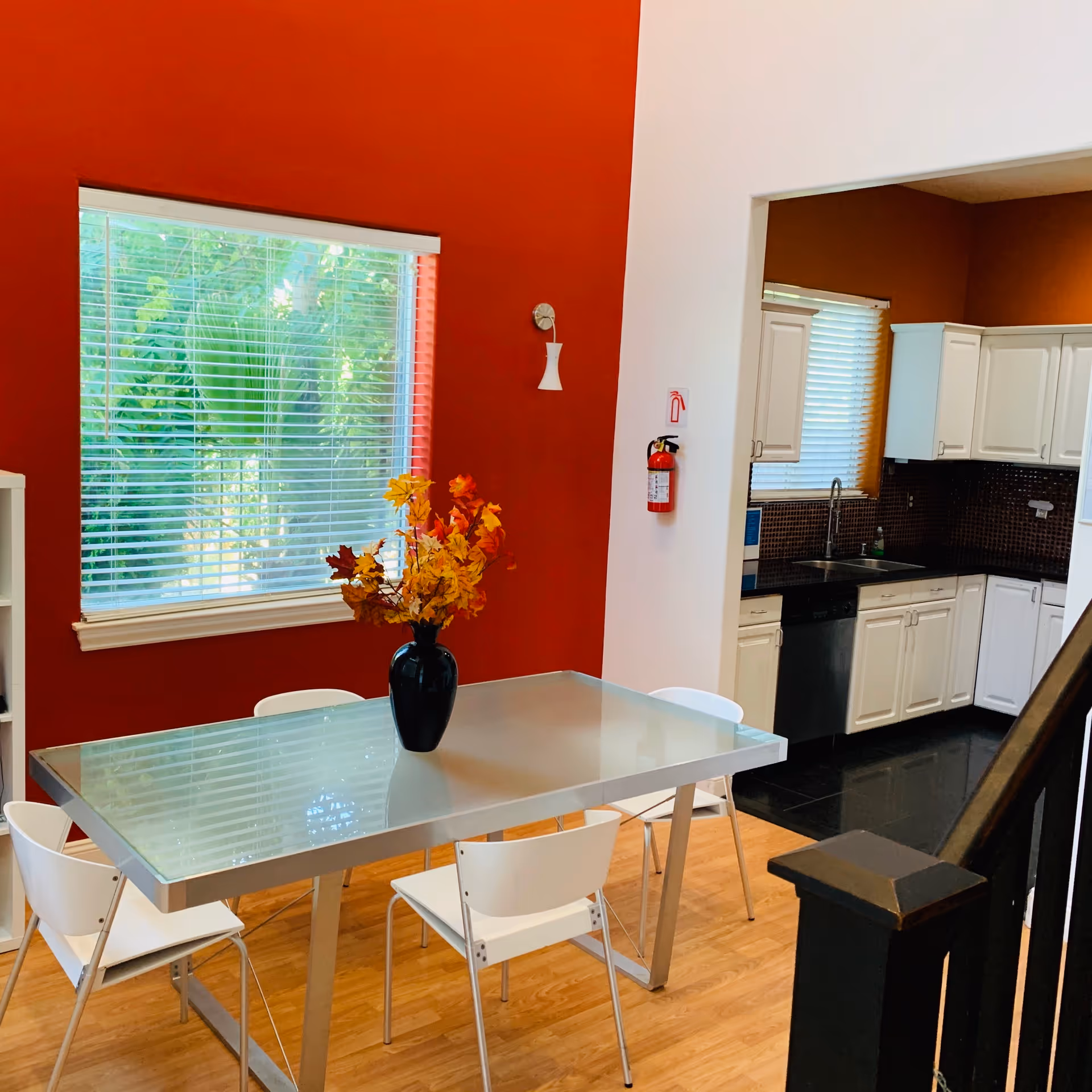 A dining area with a glass table and four white chairs on a wooden floor. A black vase with autumn-colored leaves is placed on the table. The wall behind the table is painted red with a window covered by white blinds. To the right, there is an open doorway leading to a kitchen with white cabinets, a black countertop, and a window with blinds. A fire extinguisher is mounted on the white wall near the doorway.