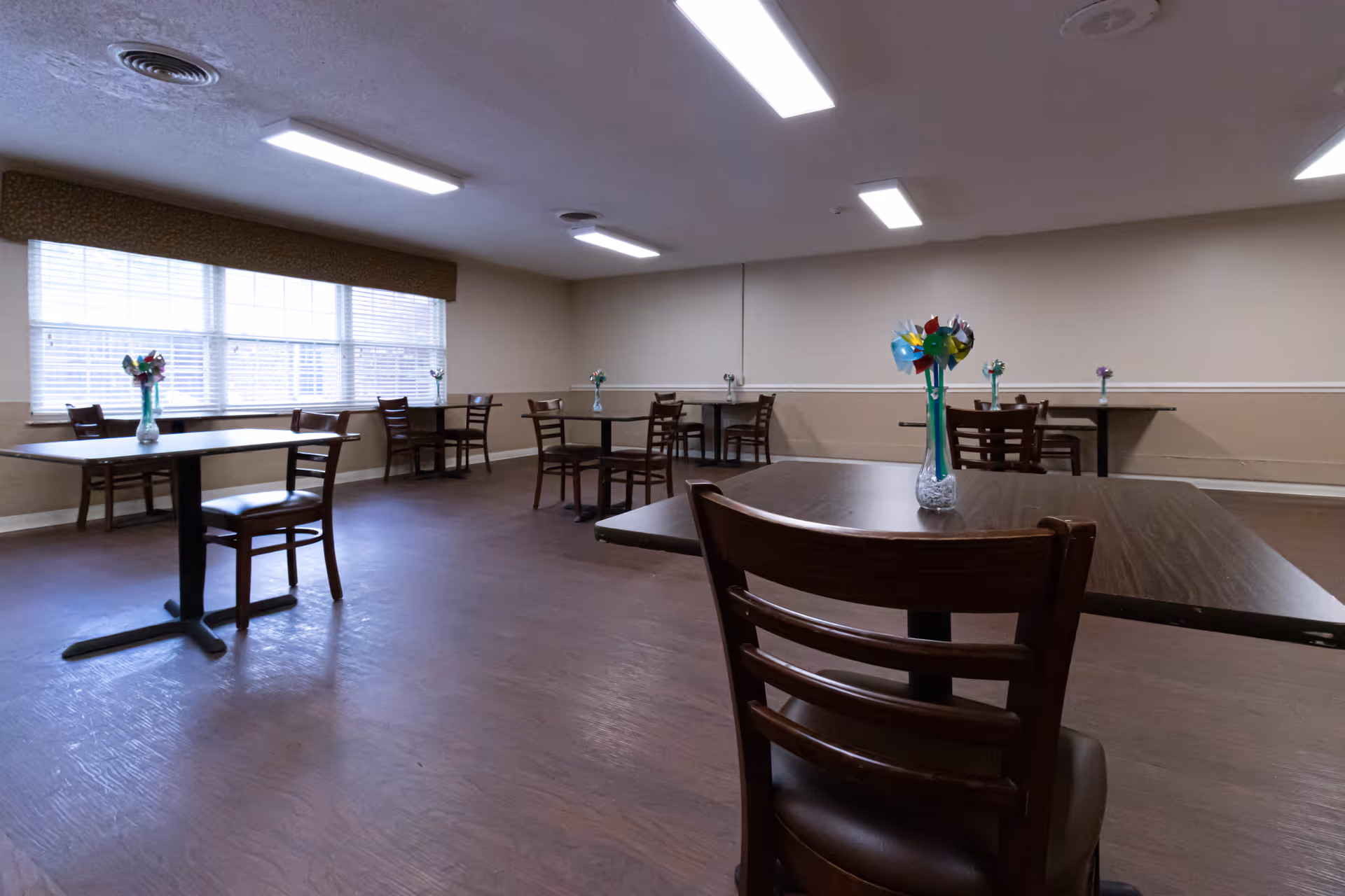 A dining room with several wooden tables and chairs arranged throughout the space. Each table has a small vase with colorful pinwheel decorations. Large windows with blinds allow natural light to enter the room. The floor is wooden, and the walls are painted beige with a white trim.