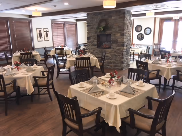 A dining room with multiple tables covered in white tablecloths, each set with folded napkins, cups, and silverware. The room features wooden chairs, a stone fireplace in the center, wooden blinds on the windows, and soft overhead lighting.