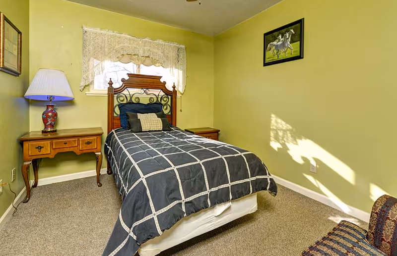 A small bedroom with a single bed featuring a wooden headboard and black and white checkered bedding. There is a wooden nightstand with a decorative lamp on the left side of the bed and another small table on the right. The walls are painted light green, and a window with lace curtains is above the bed. A framed picture of horses hangs on the wall to the right, and part of a patterned chair is visible in the bottom right corner.