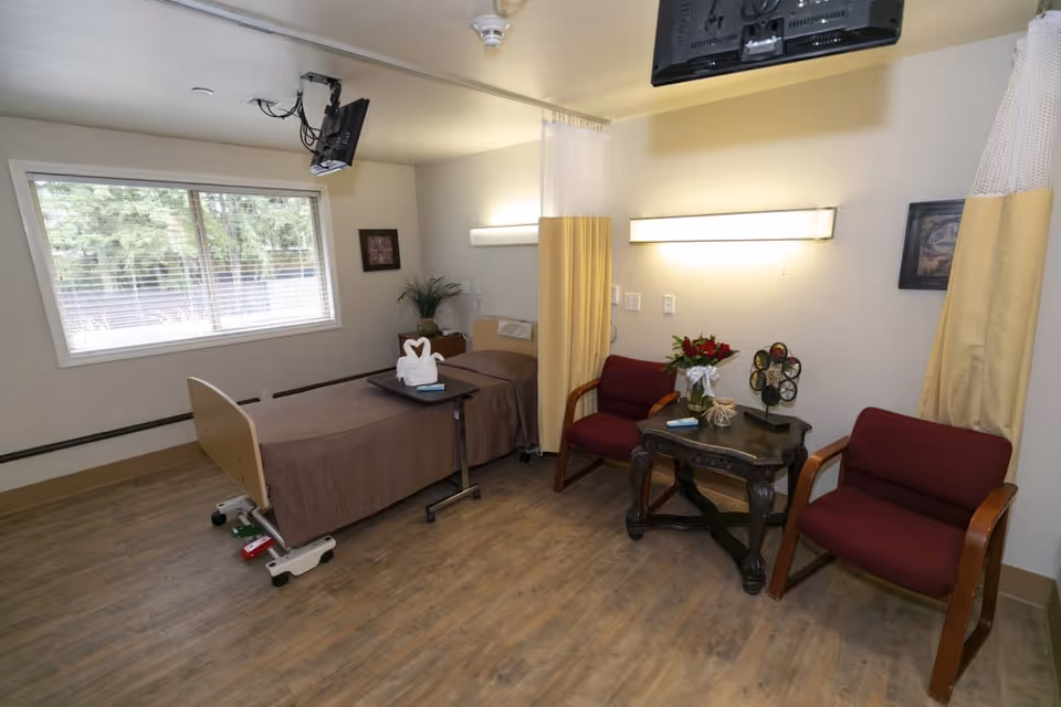 A patient room in a rehabilitation center featuring a single hospital bed with brown bedding and a towel folded into a swan shape on a tray table. The room has two maroon chairs with wooden arms, a small dark wooden table with a flower vase and decorative item, a large window with blinds, and a wall-mounted TV. The floor is wood-style, and there are light fixtures on the walls along with a privacy curtain.