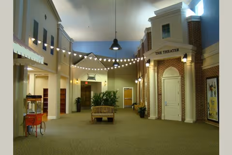 Interior hallway of a senior living facility with a high ceiling, string lights hanging across, a popcorn machine on the left, a wooden bench with plants behind it in the center, and a door labeled 'The Theater' on the right side with brick and white trim walls.
