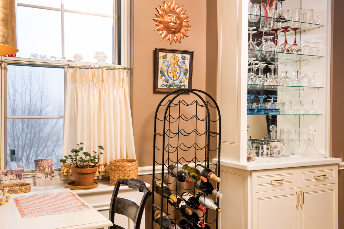 A cozy corner of a room with a window partially covered by cream-colored curtains. Below the window is a small table with a pink and white placemat, a potted plant, and some decorative items. Next to the table is a black metal wine rack holding several bottles of wine. On the wall above the wine rack, there is a decorative sun sculpture and a framed tile artwork. To the right, there is a white cabinet with glass shelves displaying various glassware including wine glasses and decanters.